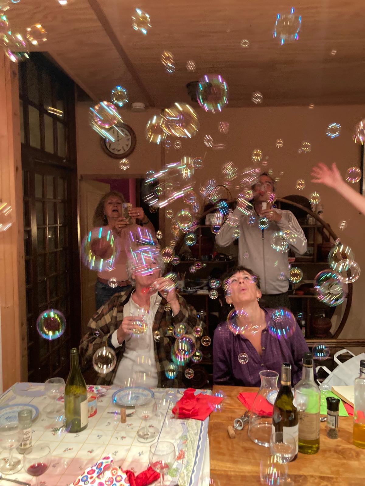 Autour d'une table dressée, personnes riantes en faisant flotter des bulles de savon dans une pièce en bois