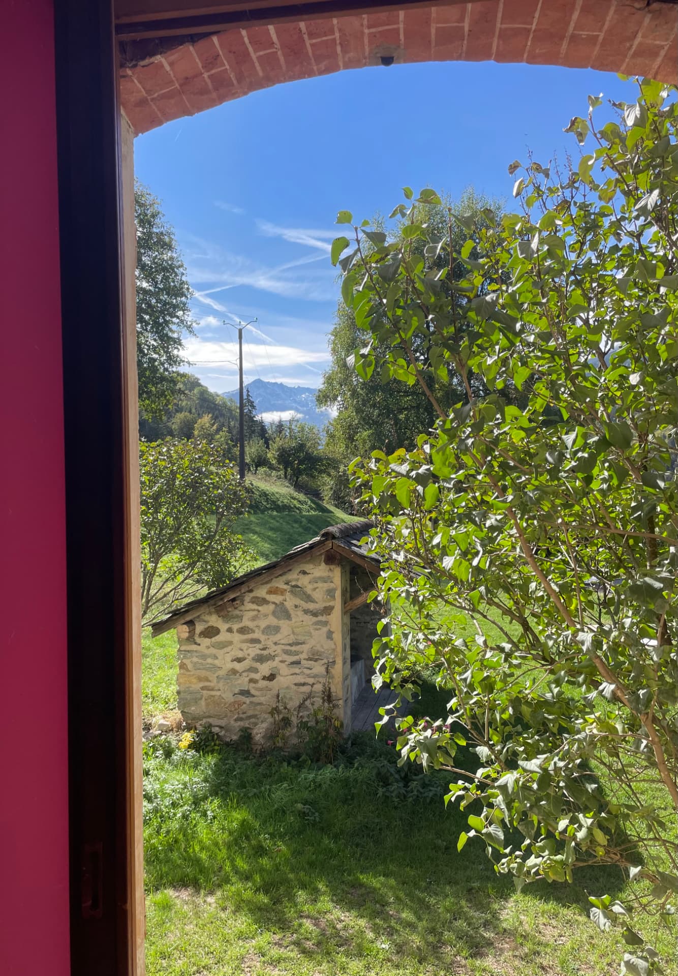 Vue depuis une fenêtre : petite cabane en pierre, jardin, arbres et montagne sous un ciel bleu